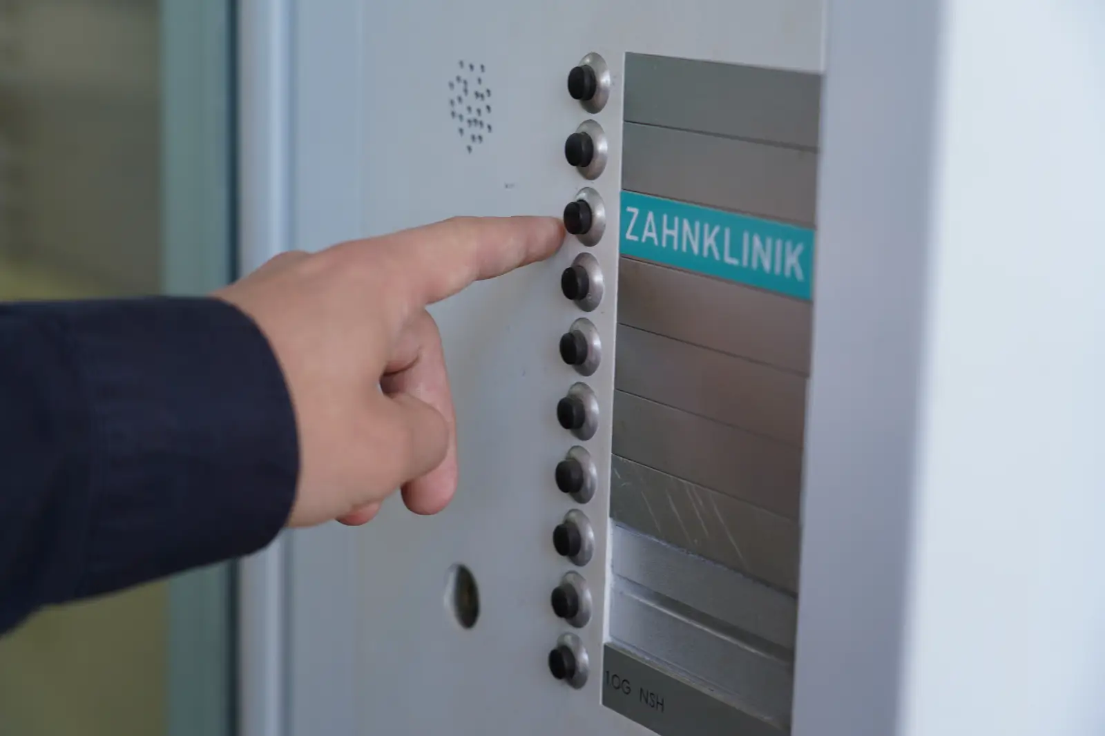 Hand drückt am Eingang die Klingel mit der Aufschrift Zahnklinik der ZBB Zahnklinik Basel